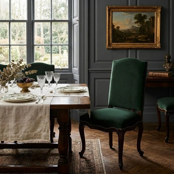 A dark green velvet dining chair with nailhead trim stands beside a wooden dining table set with plates and glassware in a classic room with dark gray paneled walls and a framed landscape painting.