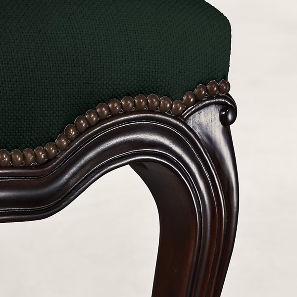 Close-up of a dark green upholstered chair or stool with a carved dark brown wooden frame and a decorative beaded trim along the seat edge.