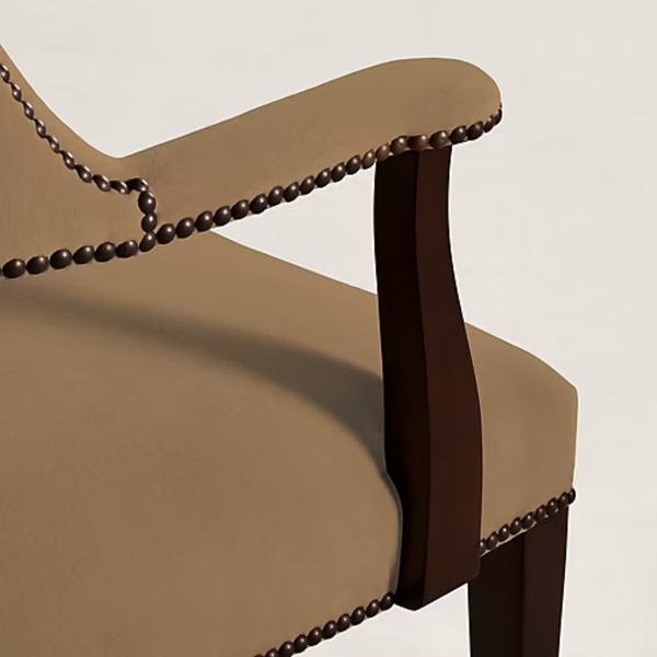 Close-up of an upholstered chair showing a warm beige fabric seat and back, dark brown wooden armrests and leg, and decorative dark metal nailhead trim along the arm and edge.