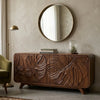 A dark brown wooden sideboard with large carved tropical leaf patterns on the front, styled in a room setting under a round gold-framed mirror.