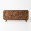 A mid-century modern wooden sideboard featuring three doors with large, hand-carved monstera leaf patterns on the front, supported by two tapered legs, shown against a plain white background.