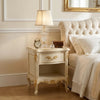 An elegant cream and gold bedside table styled next to a tufted bed in a warm, classic bedroom setting.