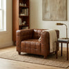 A brown tufted leather armchair in a styled living room setting, featuring a cream throw blanket draped over the side, next to a wooden side table with a brass desk lamp.