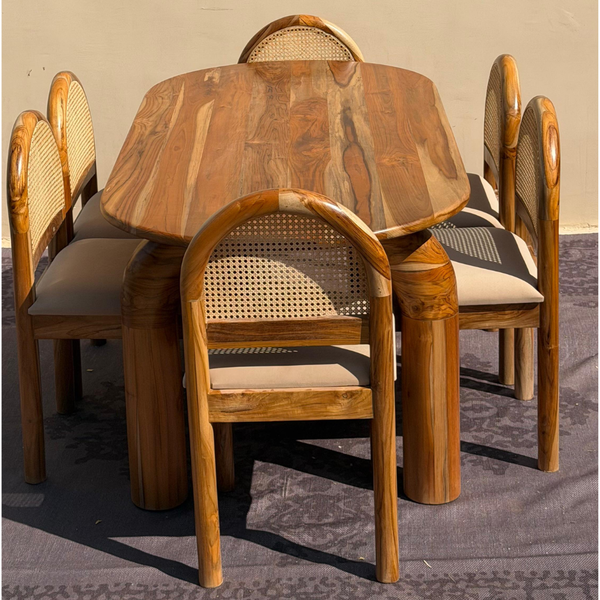 A rustic dining set featuring a round wooden table and four matching chairs with woven cane backs and light beige upholstered seats, photographed outdoors on a patterned rug.
