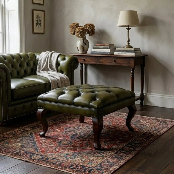 An olive green tufted leather ottoman with dark wooden legs is styled in a living room next to a matching green leather Chesterfield sofa, set on a patterned red and blue rug.