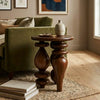 A dark brown wooden end table with ornate turned legs stands beside a green sofa in a styled living room. A small textured vase and a tray with glasses sit on top of the table, with a rug and books in the foreground.