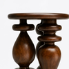 A close-up of a dark brown wooden stool or side table with a round top and turned pedestal-style legs on a light gray background.