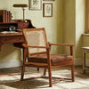 A wooden armchair with a woven cane backrest and a brown upholstered seat cushion, placed in a vintage-style room next to a wooden desk.