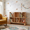 Wooden bookshelf with books and toys in a child's room with cloud-themed wallpaper.