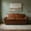 A brown leather loveseat sofa placed in a styled room with a green brick wall, a framed landscape painting above, a patterned rug underneath, and a floor lamp to the right.