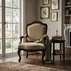 An ornate vintage-style armchair with carved dark wood and beige upholstery, placed in a decorated room beside a black side table with books and a draped green throw blanket.