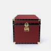 A vintage-style red leather trunk with black leather trim and brass hardware, including corner protectors and a central latch, photographed on a light gray background.