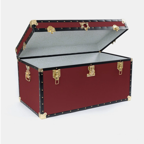 A vintage-style trunk or storage chest with a deep red exterior, black trim, brass corner and latch hardware, and a light gray interior, shown open on a plain light gray background.