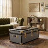 A dark gray vintage-style trunk coffee table with brass hardware and corner protectors, styled in a cozy living room setting with a green sofa, bookshelves, and a patterned rug.