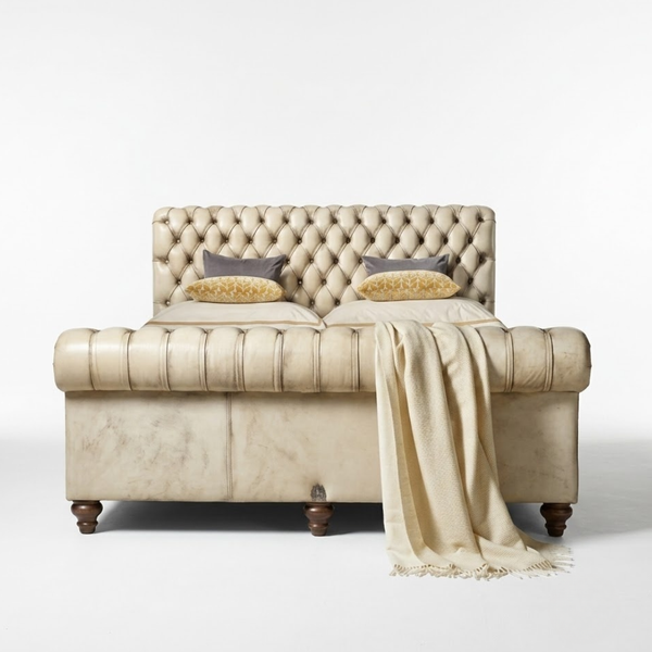 A tufted upholstered bed frame in a warm cream-beige tone, styled with two gray pillows, two gold patterned accent pillows, and a matching cream throw blanket draped over the front. The bed is photographed against a seamless white studio background.