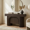A dark brown console table with ribbed legs and an arched center cabinet, styled in a warm, modern room with a round wall mirror, books, a vase, and a chair partially visible.