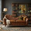 A brown leather chaise lounge sofa with tufted detailing, styled in a dark, elegant room with a floor lamp, side table, patterned rug, and bookshelves in the background.