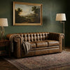 A brown leather Chesterfield sofa in a styled living room with a dark green wall, a framed landscape painting, a patterned area rug, a floor lamp, and a table lamp.