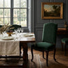 A dark green velvet dining chair with nailhead trim stands beside a wooden dining table set with plates and glassware in a classic room with dark gray paneled walls and a framed landscape painting.