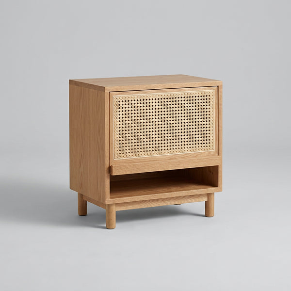 Wooden side table with wicker pattern next to a wooden chair in a room with light-colored tiles.