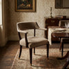A classic upholstered armchair in a warm taupe-beige velvet fabric with dark wood legs and silver nailhead trim, set in a vintage-style dining room.