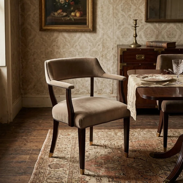 A classic upholstered armchair in a warm taupe-beige velvet fabric with dark wood legs and silver nailhead trim, set in a vintage-style dining room.