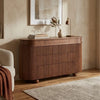A curved wooden credenza or sideboard with vertical fluted drawer fronts, shown in a styled room setting with a lamp, books, a vase, framed wall art, a chair, and a rug.