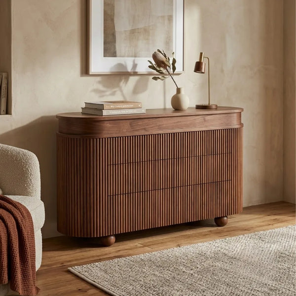 A curved wooden credenza or sideboard with vertical fluted drawer fronts, shown in a styled room setting with a lamp, books, a vase, framed wall art, a chair, and a rug.