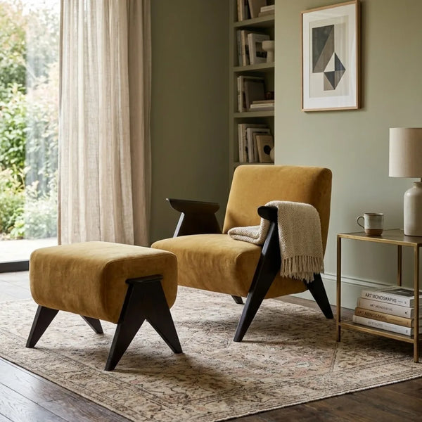 A mustard yellow velvet lounge chair with dark wood legs is styled in a living room next to a matching ottoman, a side table, and a beige throw blanket.