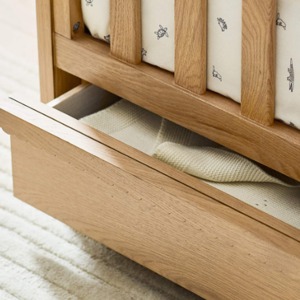 Wooden crib with open drawer revealing a mattress and fitted sheet.