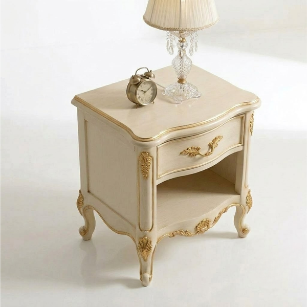 An ornate cream-colored bedside table with gold accents, featuring a single drawer, an open shelf, and curved legs. A vintage-style brass alarm clock and a table lamp with a crystal base sit on top.