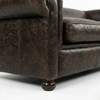 Close-up of a dark brown distressed leather sofa showing the armrest, cushion, and turned wooden leg on a white studio background.
