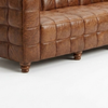 Close-up of a brown tufted leather sofa showing the armrest, seat cushion, and turned wooden legs on a white studio background.