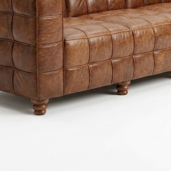Close-up of a brown tufted leather sofa showing the armrest, seat cushion, and turned wooden legs on a white studio background.