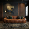 A brown leather sofa with vertical channel tufting, styled in a dark, moody living room with dark green and charcoal throw pillows, a floor lamp, a framed abstract painting, and a patterned rug.