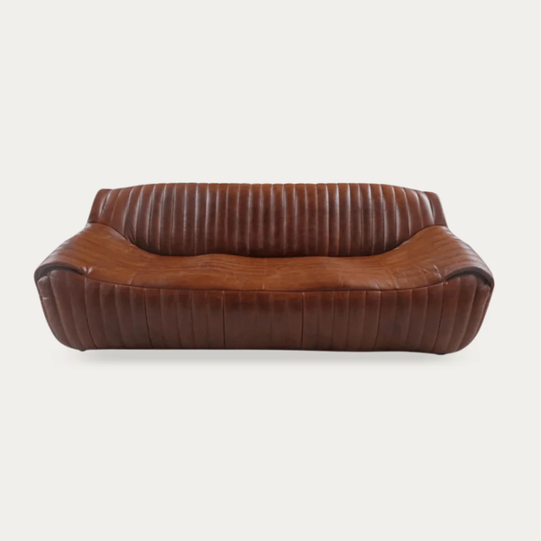 A vintage-style brown leather loveseat sofa with a curved, low-profile silhouette, featuring vertical channel tufting on the backrest and arms, shown against a plain light gray background.