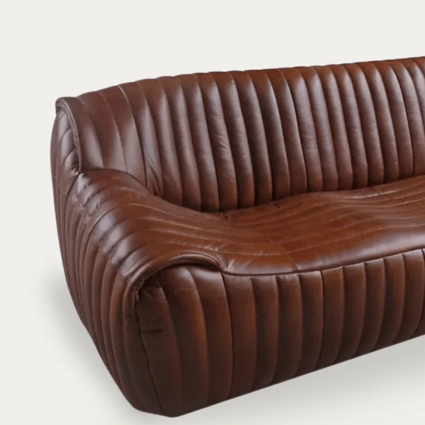 A close-up of a brown leather sofa with vertical channel tufting on the backrest and seat, shown against a light gray background.
