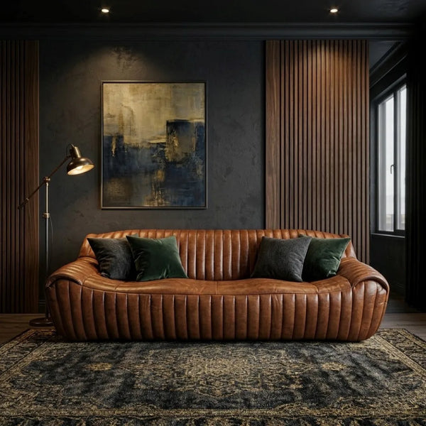 A brown leather sofa with vertical channel tufting, styled in a dark, moody living room with dark green and charcoal throw pillows, a floor lamp, a framed abstract painting, and a patterned rug.