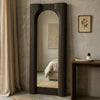 A tall, narrow full-length mirror with a dark brown fluted frame, standing in a styled bedroom setting with a concrete-textured wall, a side table, and a curtain.