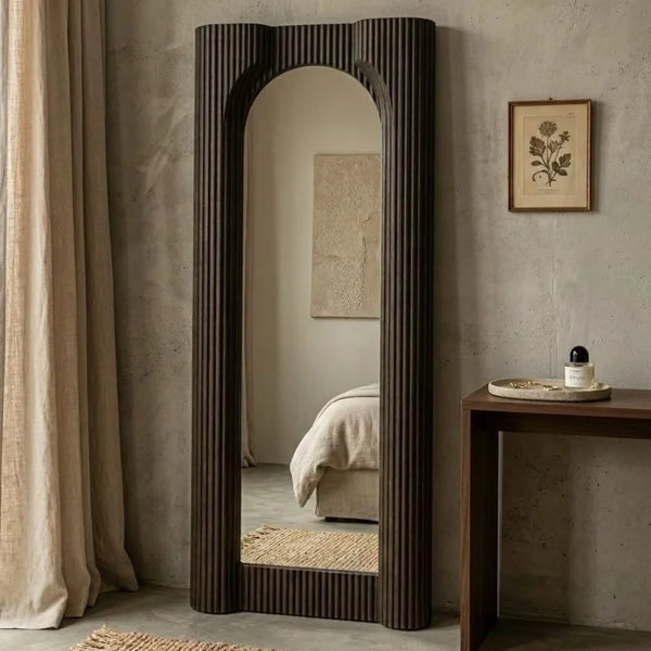 A tall, narrow full-length mirror with a dark brown fluted frame, standing in a styled bedroom setting with a concrete-textured wall, a side table, and a curtain.