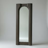 A tall, narrow arched floor mirror with a dark ribbed frame, standing on a concrete floor against a textured gray wall.