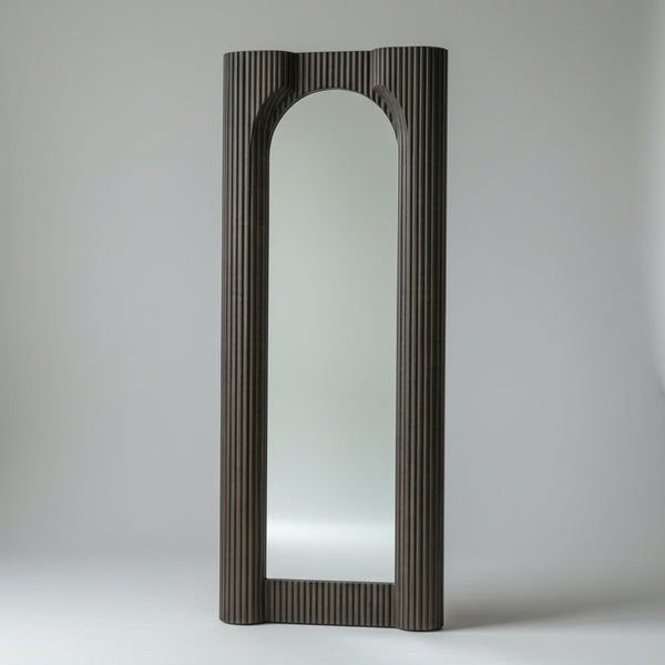 A tall, narrow arched floor mirror with a dark ribbed frame, standing on a concrete floor against a textured gray wall.