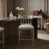 A single upholstered bar stool with a distressed silver frame and cream seat cushion, placed in a classic interior setting with a marble counter, books, and a vase of white flowers.