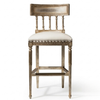 An ornate barstool with a distressed silver-painted wooden frame, featuring a spindle-back design, nailhead trim, and a cream-colored upholstered seat cushion, shown against a plain white background.