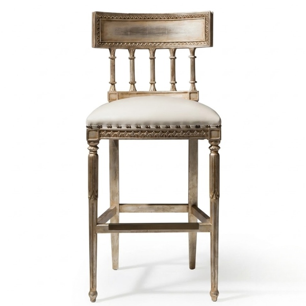 An ornate barstool with a distressed silver-painted wooden frame, featuring a spindle-back design, nailhead trim, and a cream-colored upholstered seat cushion, shown against a plain white background.