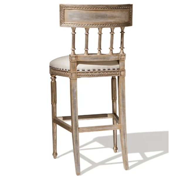 A distressed light brown wooden barstool with a cream upholstered seat, decorative nailhead trim, and a spindle-back design, shown against a plain white background.