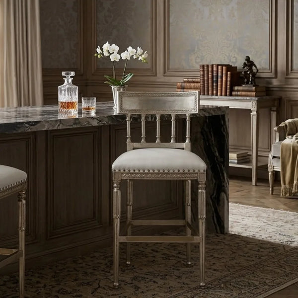 A single upholstered bar stool with a distressed silver frame and cream seat cushion, placed in a classic interior setting with a marble counter, books, and a vase of white flowers.