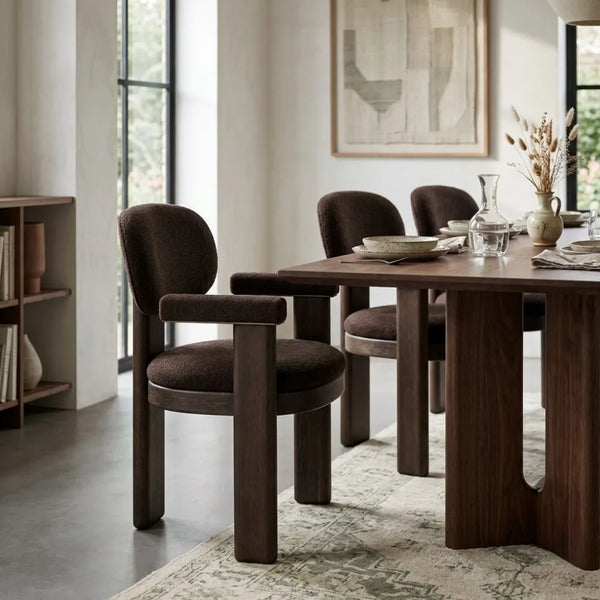 A dining room scene featuring a dark wood dining table and four upholstered armchairs in a warm brown fabric, set on a patterned area rug in a modern interior.