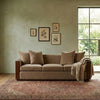 A beige upholstered sofa with dark wood arms and legs, styled with four matching throw pillows and a knitted throw blanket, set on a large patterned area rug in a living room with a textured sage green wall and a window.