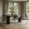 A modern dining room setup featuring a dark rectangular table with a cylindrical pedestal base, surrounded by four upholstered dining chairs. The furniture is arranged on a textured beige rug in front of large windows with beige curtains, looking out onto a green garden.